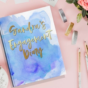 May include: A hardcover diary with a watercolor design in shades of blue and purple. The gold foil text reads "Sandra's Engagement Diary". Decorative tape, pencils, and flowers are arranged around the diary on a pink surface.