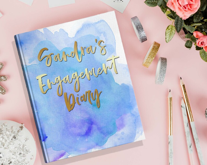 May include: A hardcover diary with a watercolor design in shades of blue and purple. The gold foil text reads "Sandra's Engagement Diary". Decorative tape, pencils, and flowers are arranged around the diary on a pink surface.