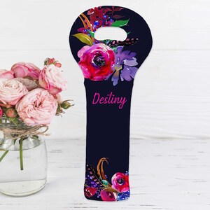 May include: A navy blue wine bottle bag with a floral design featuring pink and purple flowers, green leaves, and feathers. The word "Destiny" is written in pink script. A bouquet of pink roses in a glass jar is in the background.