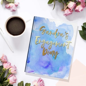 May include: A hardcover diary with the text "Sandra's Engagement Diary" in gold lettering. The cover features a watercolor design in shades of blue and purple. A white pen and a cup of coffee are next to the diary, surrounded by pink roses.