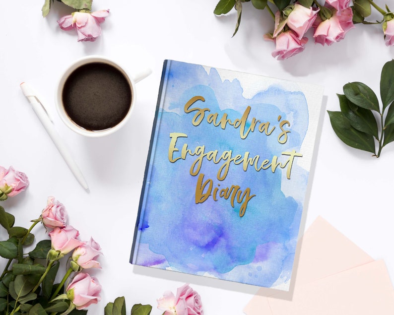 May include: A hardcover diary with the text "Sandra's Engagement Diary" in gold lettering. The cover features a watercolor design in shades of blue and purple. A white pen and a cup of coffee are next to the diary, surrounded by pink roses.