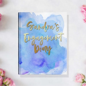 May include: A hardcover diary with a watercolor design in shades of blue and purple. The gold text reads "Sandra's Engagement Diary." Pink roses are arranged around the diary on a white surface.