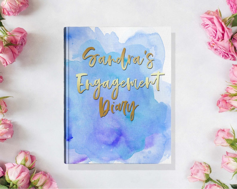 May include: A hardcover diary with a watercolor design in shades of blue and purple. The gold text reads "Sandra's Engagement Diary." Pink roses are arranged around the diary on a white surface.