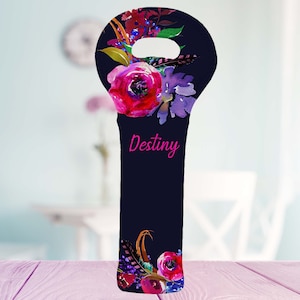 May include: A navy blue wine bottle bag with a floral design featuring pink and purple flowers, berries, and feathers. The word "Destiny" is written in pink script. The bag has a handle at the top.