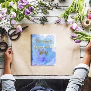 May include: A diary with the text "Sandra's Engagement Diary" in gold lettering on a blue watercolor background. The diary is surrounded by flowers, scissors, and other craft supplies on a wooden surface.