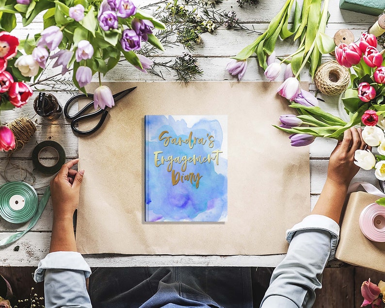 May include: A diary with the text "Sandra's Engagement Diary" in gold lettering on a blue watercolor background. The diary is surrounded by flowers, scissors, and other craft supplies on a wooden surface.