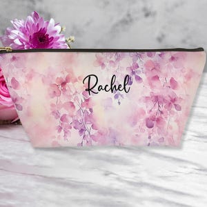 May include: A pink floral makeup bag with the name "Rachel" in black script. The bag features a watercolor design of pink and purple flowers. It has a black zipper and a gold-colored pull.