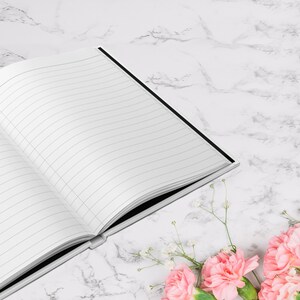 May include: An open, lined notebook with a black spine rests on a white marble surface. Pink carnations and small white flowers are arranged in the lower right corner. The notebook's pages are blank and ready for writing.