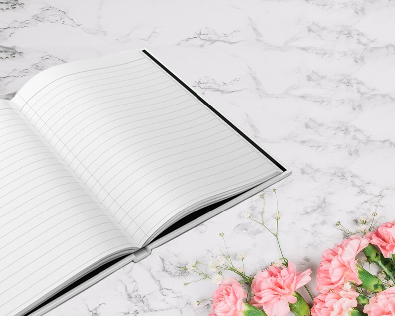 May include: An open, lined notebook with a black spine rests on a white marble surface. Pink carnations and small white flowers are arranged in the lower right corner. The notebook's pages are blank and ready for writing.