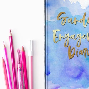 May include: A pink and magenta stationery set next to a diary. The diary cover has a watercolor design in blue and purple with gold lettering that reads "Sandra's Engagement Diary". Pencils and a pen are arranged on a white surface.