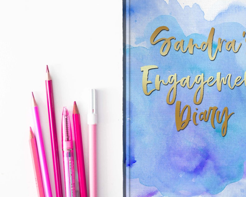 May include: A pink and magenta stationery set next to a diary. The diary cover has a watercolor design in blue and purple with gold lettering that reads "Sandra's Engagement Diary". Pencils and a pen are arranged on a white surface.