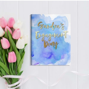 May include: A diary with a blue watercolor design and gold lettering that reads "Sandra's Engagement Diary." A bouquet of pink and white tulips with green stems and leaves is next to the diary on a white surface.