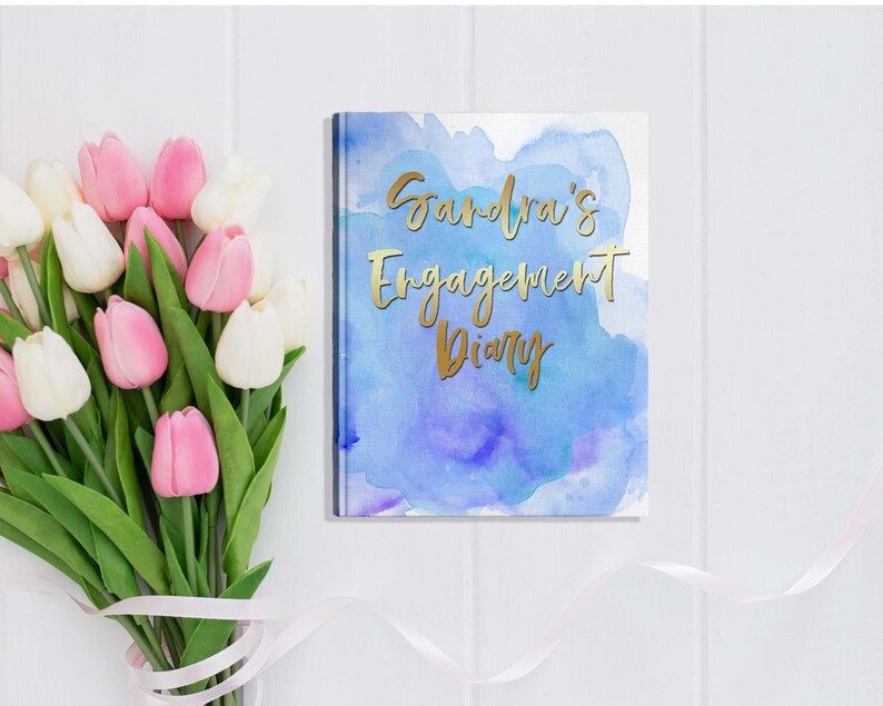 May include: A diary with a blue watercolor design and gold lettering that reads "Sandra's Engagement Diary." A bouquet of pink and white tulips with green stems and leaves is next to the diary on a white surface.
