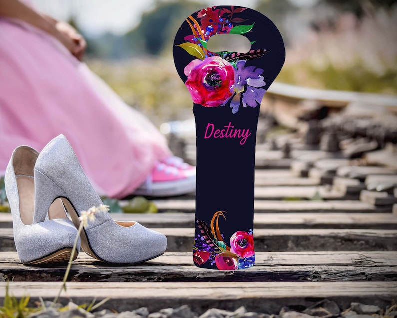 May include: A navy blue wine bottle bag with a floral design and the word "Destiny" in pink. The bag features red, purple, and blue flowers. Silver glitter high heels are in the foreground.