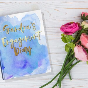 May include: A hardcover diary with the text "Sandra's Engagement Diary" in gold lettering. The cover features a watercolor design in shades of blue and purple. Pink and red roses are arranged on a white wooden surface.