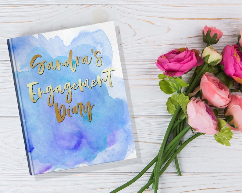 May include: A hardcover diary with the text "Sandra's Engagement Diary" in gold lettering. The cover features a watercolor design in shades of blue and purple. Pink and red roses are arranged on a white wooden surface.