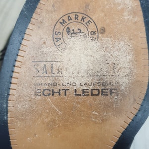 May include: Close-up of the sole of a black leather shoe. The sole is brown and has a stitched edge. The sole is marked with the text "SALZ MARKETER" and "HAND- UND LAUFSCHUH ECHT LEDER".