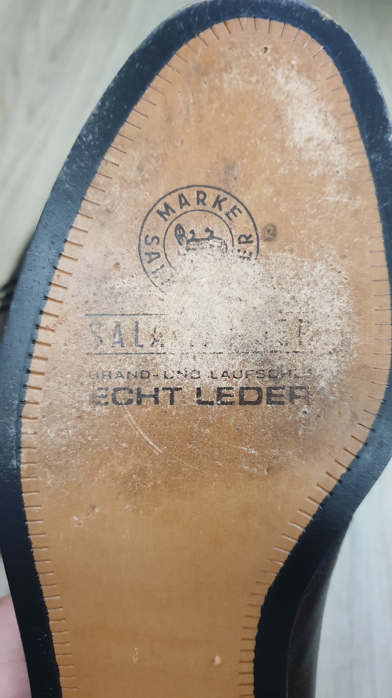 May include: Close-up of the sole of a black leather shoe. The sole is brown and has a stitched edge. The sole is marked with the text "SALZ MARKETER" and "HAND- UND LAUFSCHUH ECHT LEDER".