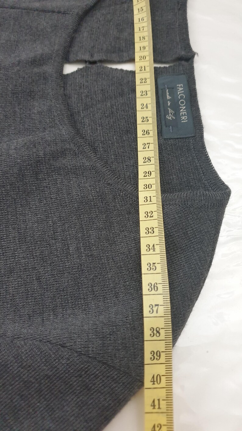 Puede incluir: Primer plano de un su&eacute;ter gris oscuro con una cinta m&eacute;trica al lado. El su&eacute;ter tiene una etiqueta que dice "FALCONERI made in Italy". La cinta m&eacute;trica muestra la longitud del su&eacute;ter en cent&iacute;metros.