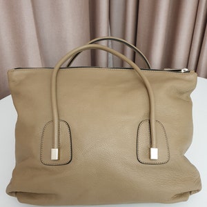 May include: A large, light brown leather tote bag with two top handles and a zipper closure. The bag has a textured, pebbled leather finish.