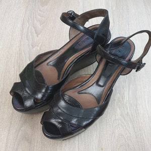 Puede incluir: Un par de sandalias de cuña de cuero negro con una correa de tobillo y puntera abierta.