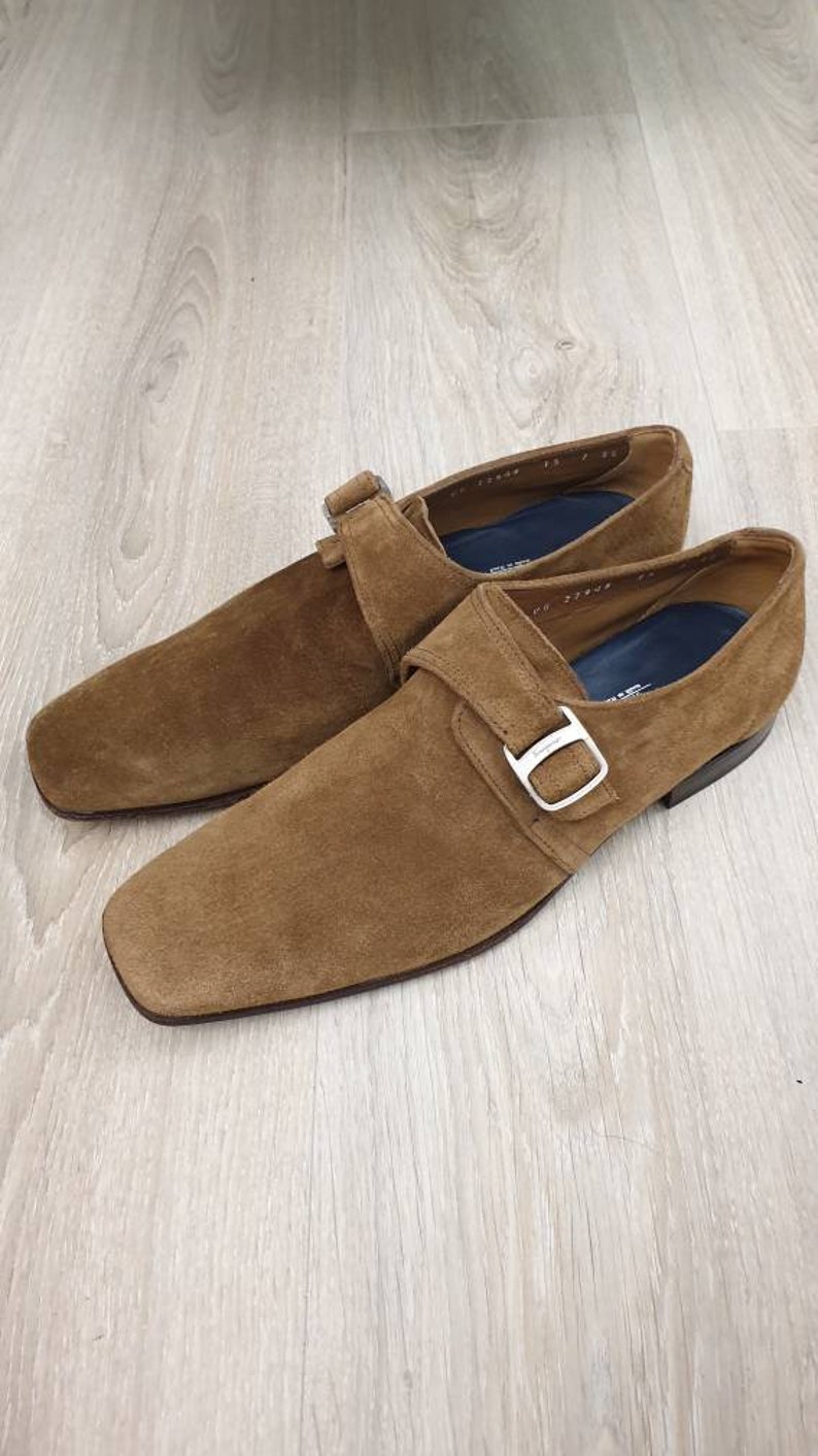 May include: A pair of brown suede monk strap shoes with a silver buckle. The shoes have a square toe and a low heel.