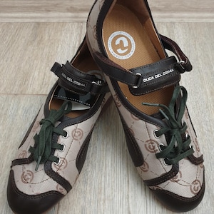 Op de afbeelding: Paar bruine en beige Duca Del Cosma schoenen. De schoenen hebben een herhalend patroon en donkerbruine accenten. Ze hebben groene veters, een bandsluiting en de merknaam op de band.