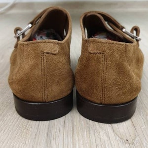 May include: A pair of brown suede shoes with buckle fastenings. The shoes have a dark brown sole and are made of a soft, textured material.