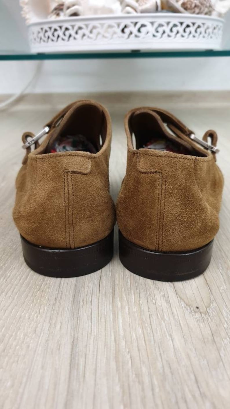 May include: A pair of brown suede shoes with buckle fastenings. The shoes have a dark brown sole and are made of a soft, textured material.