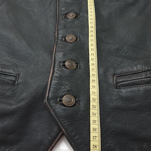 Può includere: Gilet in pelle nera con quattro bottoni in ottone sul davanti e una tasca sul lato destro. Un metro a nastro è visibile sul lato destro dell'immagine.