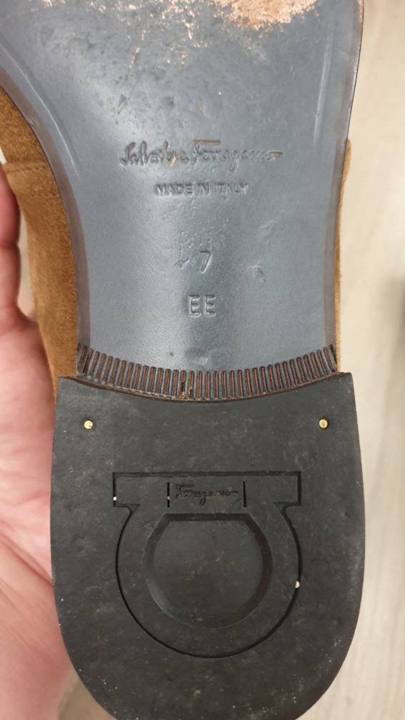 May include: Close-up of the bottom of a brown suede shoe with a black leather sole. The shoe is labelled "Salvatore Ferragamo" and "Made in Italy". The size is marked as "7 EE".