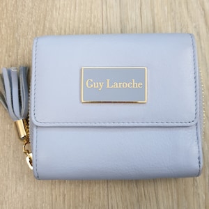 Könnte beinhalten: Eine hellblaue Lederbörse mit einem goldenen Reißverschluss und einer goldenen Plakette mit der Aufschrift "Guy Laroche".