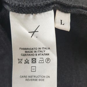 Puede incluir: Primer plano de una etiqueta de prenda negra con texto blanco. La etiqueta dice "FABBRICATO IN ITALIA MADE IN ITALY СДЕЛАНО В ИТАЛИИ" e incluye s&iacute;mbolos de cuidado. Una etiqueta blanca con la letra "L" es visible.