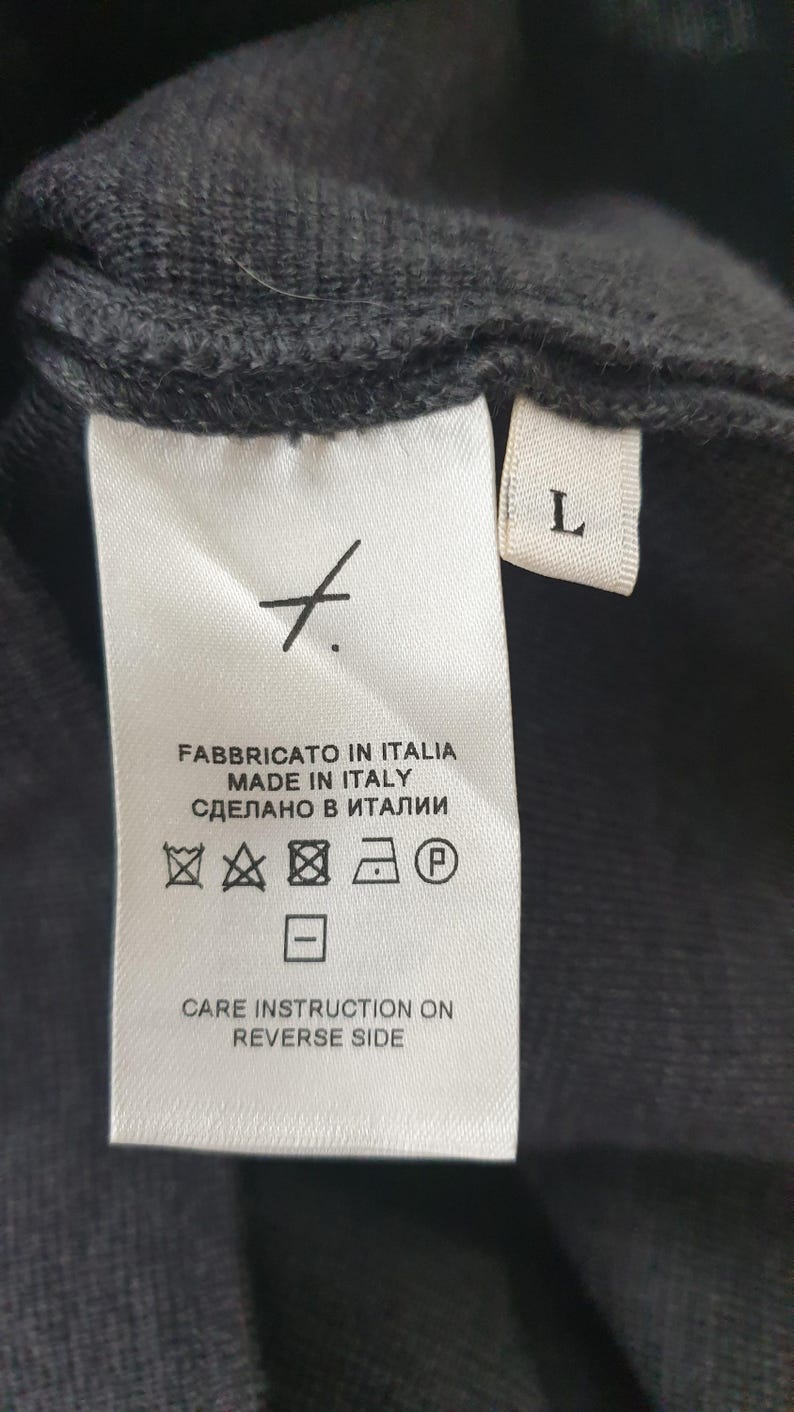 Puede incluir: Primer plano de una etiqueta de prenda negra con texto blanco. La etiqueta dice "FABBRICATO IN ITALIA MADE IN ITALY СДЕЛАНО В ИТАЛИИ" e incluye s&iacute;mbolos de cuidado. Una etiqueta blanca con la letra "L" es visible.