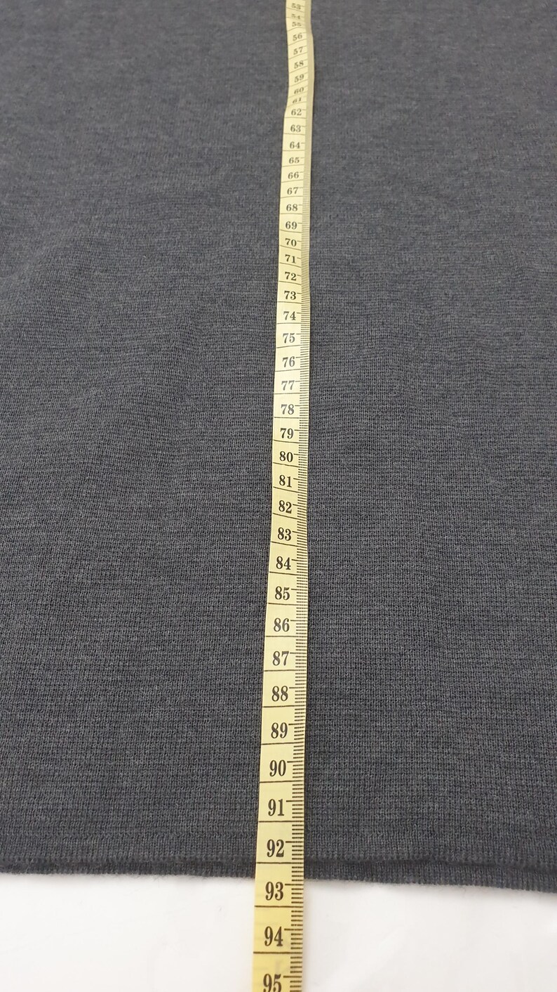 Puede incluir: Una tela gris oscuro se muestra con una cinta m&eacute;trica amarilla. La cinta est&aacute; marcada en cent&iacute;metros, con n&uacute;meros del 63 al 95. La tela es de color gris oscuro liso.