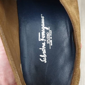 May include: Brown suede leather shoes with a buckle fastening. The inside of the shoe has a blue insole with the text "Salvatore Ferragamo STUDIO MADE IN ITALY".