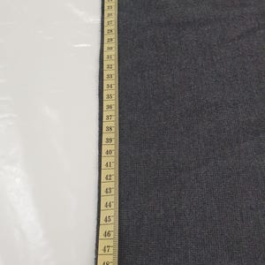 Puede incluir: Una muestra de tela gris oscuro junto a una cinta m&eacute;trica. La cinta muestra medidas de hasta 135 cm. La tela parece ser de color liso y tiene una superficie ligeramente texturizada.