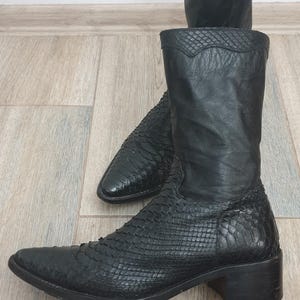 Könnte beinhalten: Schwarze Cowboy-Stiefel aus Leder mit einem strukturierten Schlangenmuster. Die Stiefel haben eine spitze Zehenpartie, einen mittelhohen Absatz und einen hohen Schaft. Der zweite Stiefel ist im Hintergrund.