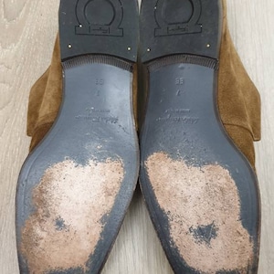 May include: A pair of brown suede leather shoes with a black leather sole. The shoes have a "Salvatore Ferragamo" logo on the sole.