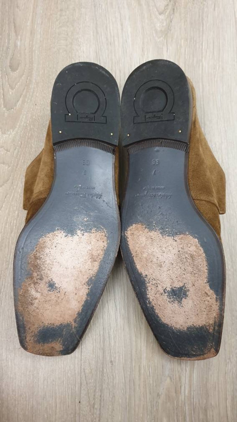 May include: A pair of brown suede leather shoes with a black leather sole. The shoes have a "Salvatore Ferragamo" logo on the sole.