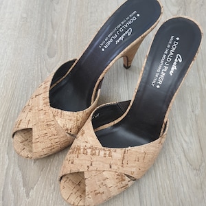 Puede incluir: Un par de sandalias de tacón de corcho con puntera abierta y plantilla de cuero negro. Las sandalias están etiquetadas "Couture Donald J Pliner Hecho en las montañas de Italia."