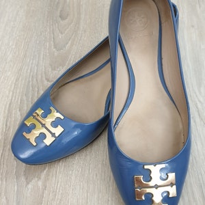 Puede incluir: Un par de bailarinas de charol azul con detalles dorados del logotipo. Los zapatos tienen una puntera redondeada y un tacón bajo. El logotipo es un diseño de doble T dorado.