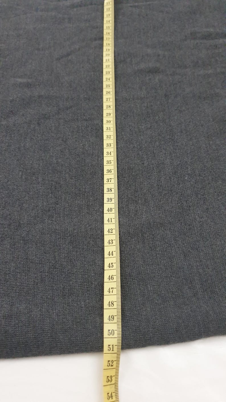 Puede incluir: Primer plano de una tela gris con una cinta m&eacute;trica amarilla. La cinta est&aacute; marcada con n&uacute;meros de 33 a 137 cm, proporcionando una referencia visual de longitud y medici&oacute;n.