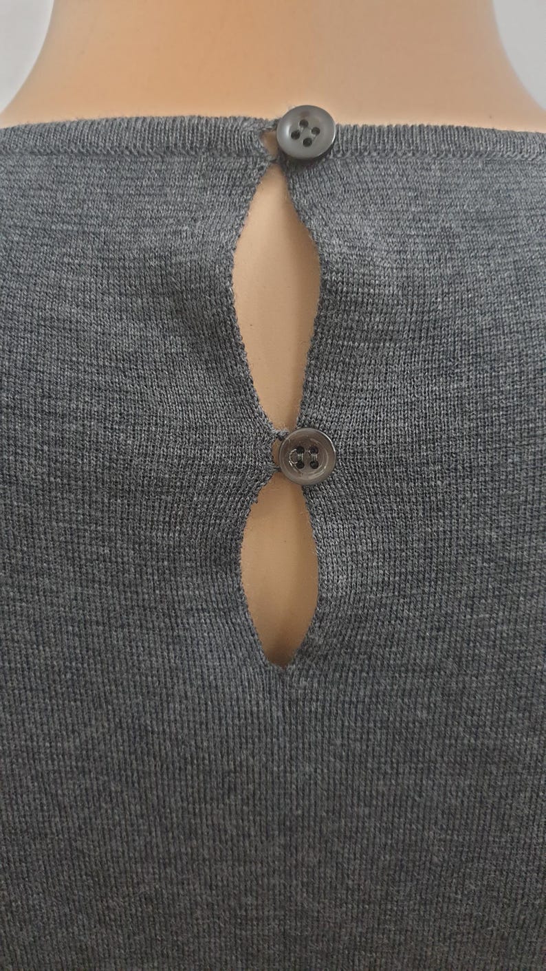 Puede incluir: Primer plano de un top de punto gris con detalle de espalda con abertura. El top presenta dos peque&ntilde;os botones redondos grises en el escote y en la mitad de la espalda. La tela tiene una textura acanalada.