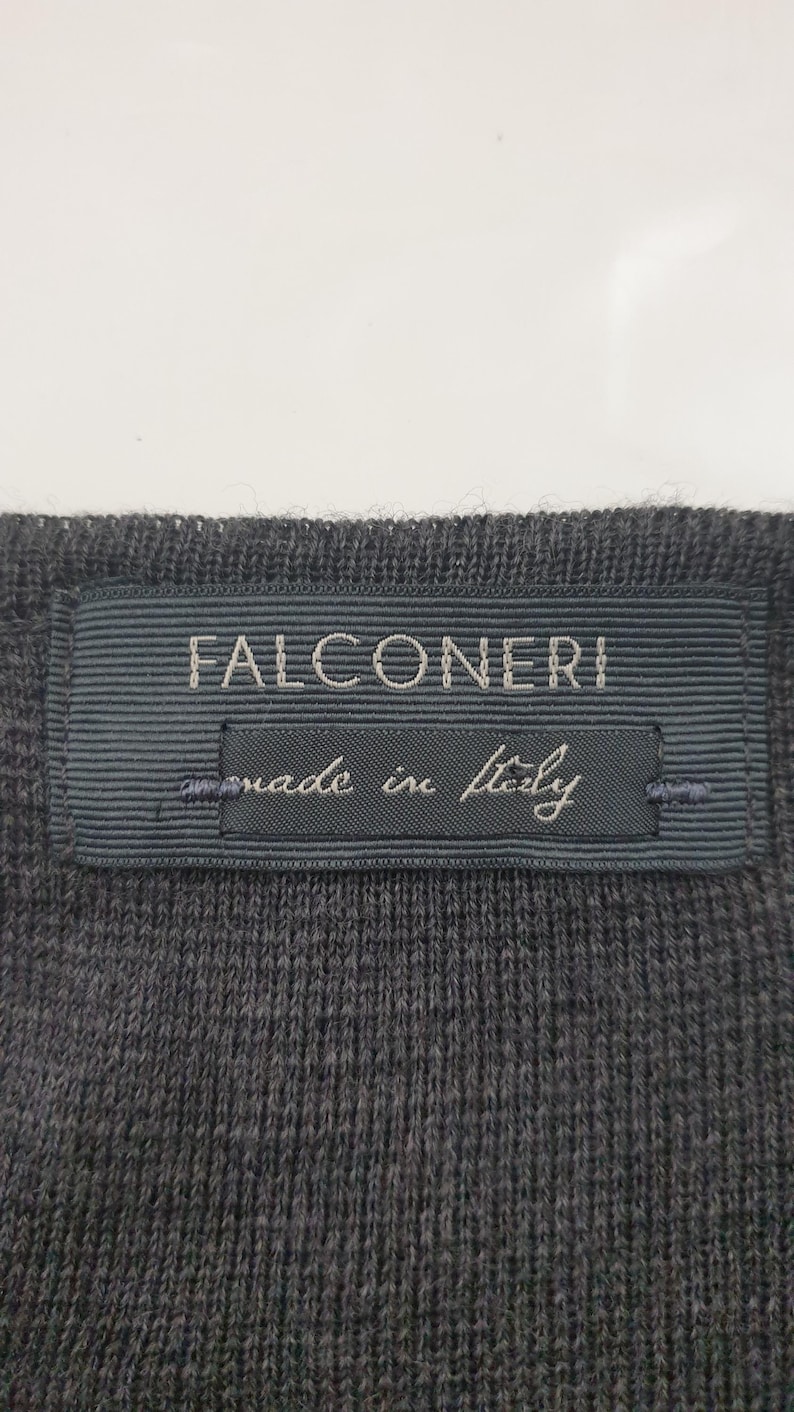 Puede incluir: Primer plano de una tela de punto gris oscuro con una etiqueta rectangular. La etiqueta, en un tono de gris m&aacute;s oscuro, dice "FALCONERI" en may&uacute;sculas, con "made in Italy" debajo.