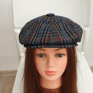 May include: A blue, gray, and red plaid newsboy cap with a black band around the bottom. The cap is on a mannequin head with long brown hair.