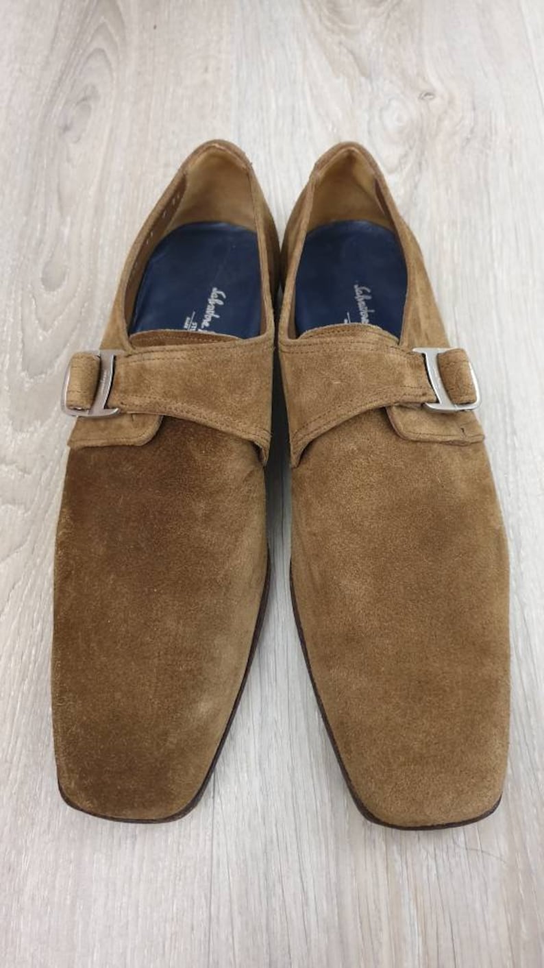 May include: A pair of brown suede monk strap shoes with silver buckles. The shoes have a square toe and a low heel.