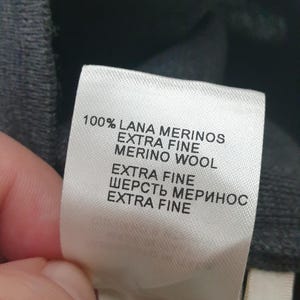 Puede incluir: Primer plano de una etiqueta de ropa con texto en varios idiomas. La etiqueta dice "100% LANA MERINOS EXTRA FINE MERINO WOOL" e incluye instrucciones de cuidado. La etiqueta es blanca con texto negro, y la prenda es gris oscuro.
