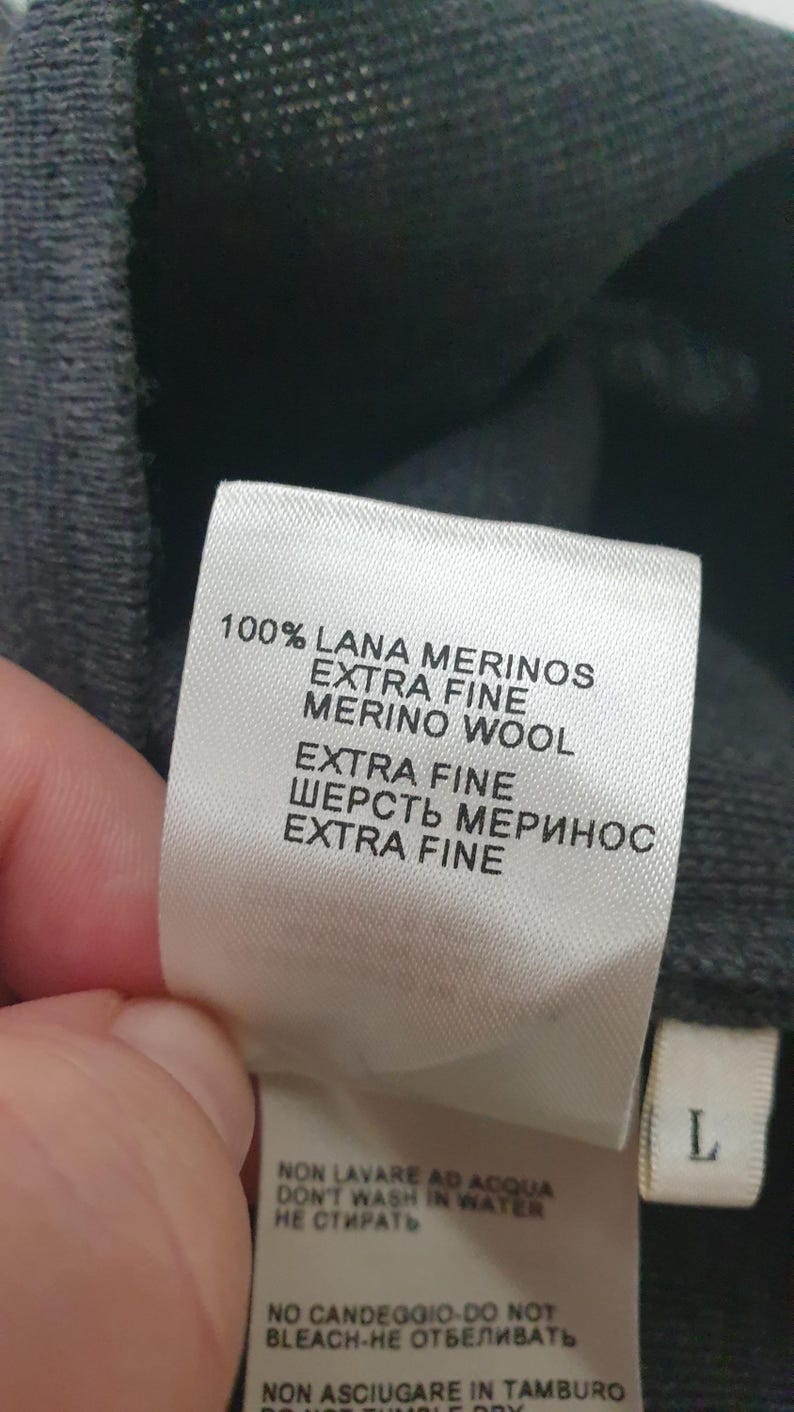 Puede incluir: Primer plano de una etiqueta de ropa con texto en varios idiomas. La etiqueta dice "100% LANA MERINOS EXTRA FINE MERINO WOOL" e incluye instrucciones de cuidado. La etiqueta es blanca con texto negro, y la prenda es gris oscuro.