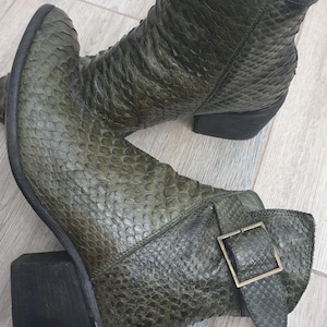 May include: A pair of olive green ankle boots featuring a textured snakeskin-like pattern, a block heel, and a side strap with a metallic buckle. The boots have a pointed toe design and a visible interior lining, showcasing a structured silhouette.